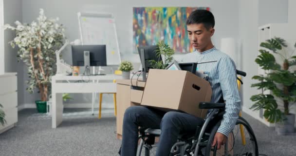 Hombre ambicioso discapacitado en camisa con belleza asiática coreana se sienta en silla de ruedas en el escritorio de la empresa en las rodillas sosteniendo caja de cartón con cosas empaquetadas, es despedido del trabajo incapaz de cumplir con sus deberes — Vídeo de stock