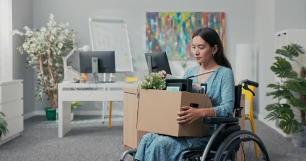 Una mujer atractiva discapacitada en vestido de belleza coreana asiática se sienta en silla de ruedas en el escritorio corporativo en su regazo sosteniendo una caja de cartón de pertenencias empacadas, cambiando de posición, siendo promovida, sonriendo — Vídeo de stock