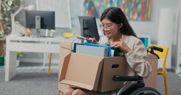 Niepełnosprawna kobieta w okularach i azjatyckiej koreańskiej urodzie siedzi na wózku inwalidzkim w biurze korporacyjnym na kolanach trzymając kartonowe pudełko zapakowanych akcesoriów, zmieniając pozycję, awansując — Wideo stockowe