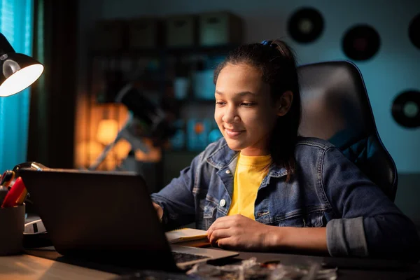 Enfocado adolescente sonriente utiliza el ordenador portátil en casa, mirando a la pantalla, charlando, leyendo o escribiendo correo electrónico, papel para la escuela Fotos De Stock Sin Royalties Gratis