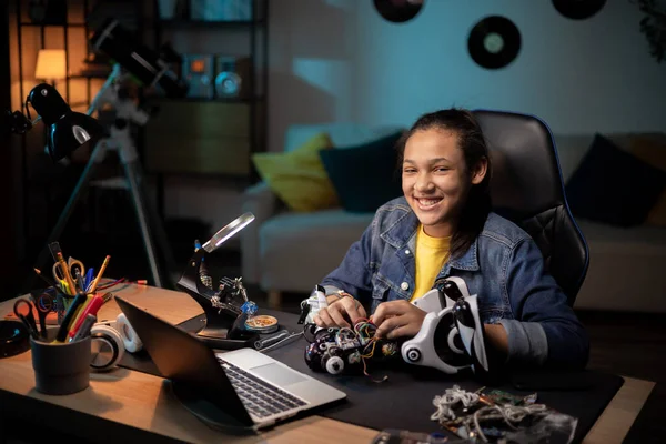 Een lachend jong meisje zit 's avonds in de kamer met weinig licht aan, tiener is geïnteresseerd in wetenschapsgebieden, robotica, assembleert haar eigen robot, verbindt de elektronica die het speelgoed van stroom voorziet Stockfoto