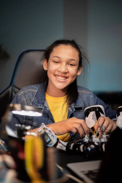 Un adolescente desarrolla una pasión por la robótica. La chica sonríe mientras se sienta en el escritorio con el ordenador portátil jugando con la electrónica que alimenta el juguete, conecta los cables de reparación del robot. Imagen De Stock