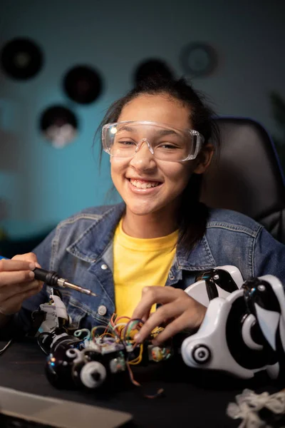 Een glimlachend getalenteerd meisje tinkelt in haar kamer bij een robot. Ze heeft een veiligheidsbril op, gebruikt een soldeerbout om elektronica in speelgoed te repareren. Stockfoto