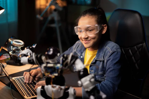 Retrato de niña sentada en el escritorio por la noche, terminando robot de soldadura que es el proyecto de la escuela, el niño está viendo tutoriales, instrucciones en el ordenador portátil, aprendiendo paso a paso cómo arreglar la electrónica Fotos De Stock Sin Royalties Gratis