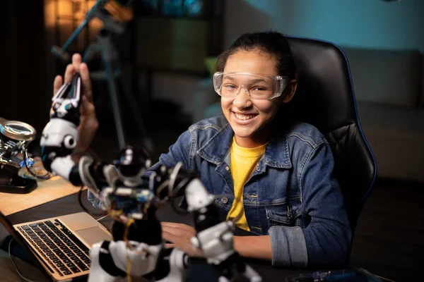 Portret van een lachend meisje dat 's avonds in de kamer aan het bureau zit, speelgoedinstructies op de laptop bekijkt, webpagina' s doorbladert, elektronicakabels van een robot trekt, repareert Stockfoto