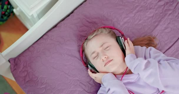 Adolescente chante avec les yeux fermés couché sur son lit dans la chambre à coucher portant des écouteurs — Video