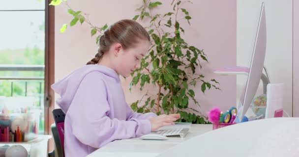 십 대 소녀가 키보드로 타자를 치고 마우스를 스크롤하고 컴퓨터로 일하고 모니터 앞에서, 자기 방에서 보라색 배경에 푸른 식물들이 있는 모습 — 비디오