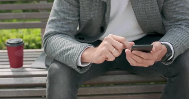 Primer plano de las manos de un hombre sentado en un banco, desplazando el teléfono inteligente, con taza de papel de café en su lado izquierdo — Vídeos de Stock