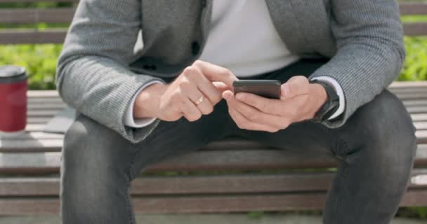 Großaufnahme der Hände eines Mannes, der auf einer Bank sitzt und mit dem Smartphone blättert, mit einer Tasse Kaffee auf der linken Seite — Stockvideo