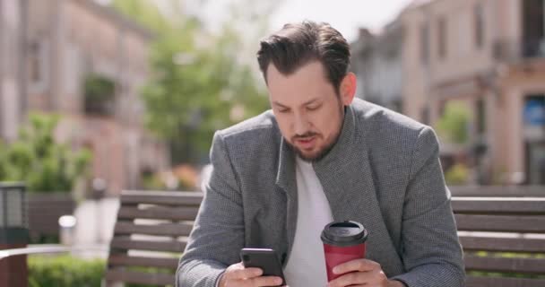 Joven está sentado en un banco en la plaza, leyendo los mensajes en el teléfono inteligente en una mano y sosteniendo el café en otro edificio en el fondo — Vídeos de Stock