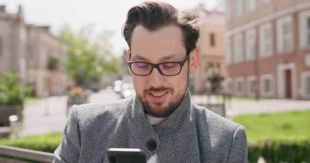 Joven, con gafas, bigotes y barba, sentado en un banco de la plaza, leyendo noticias en el smartphone Edificios de fondo — Vídeos de Stock