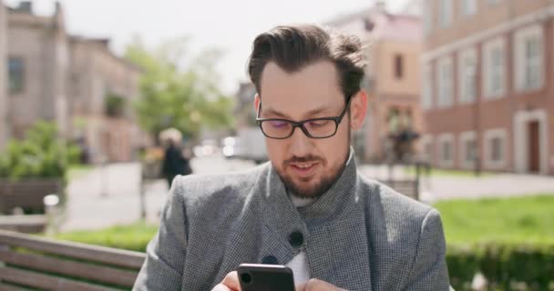 Joven, con gafas, bigotes y barba, sentado en un banco de la plaza, leyendo noticias en el smartphone Edificios de fondo — Vídeos de Stock