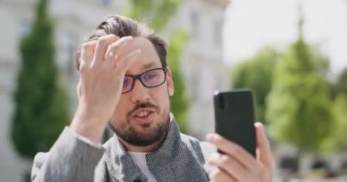 Gözlüklü, bıyıklı, sakallı, saçları düzleştiren, arka plandaki akıllı telefon binaları aracılığıyla sohbet eden genç bir adam.