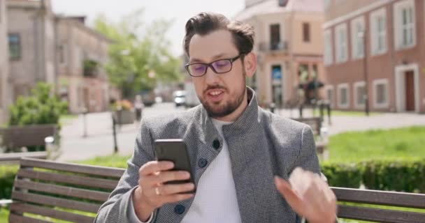 Joven, usando gafas, con bigotes y barba está sentado en un banco en la plaza, charlando a través de teléfono inteligente y saludando a mano Edificios en el fondo — Vídeos de Stock