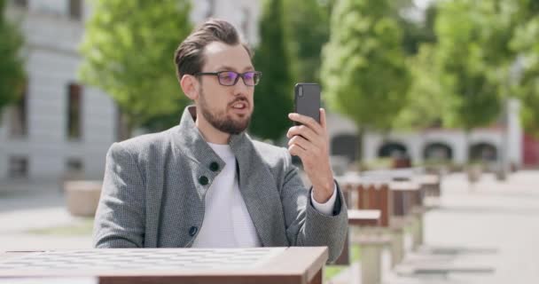 Jovem, vestindo óculos, com bigodes e barba, está sentado à mesa na praça, conversando via smartphone Edifícios em segundo plano — Vídeo de Stock