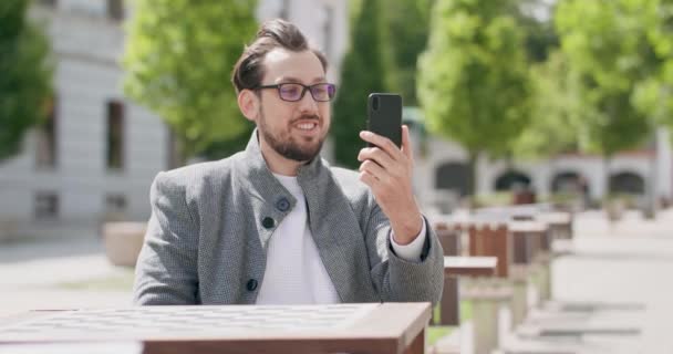 Молодий чоловік, одягнений в окуляри, з вусами і бородою, сидить за столом на площі, спілкуючись через смартфон Будівлі на фоні — стокове відео