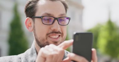 Gözlüklü, bıyıklı, sakallı, saçları düzleştiren, arka plandaki akıllı telefon binaları aracılığıyla sohbet eden genç bir adam.