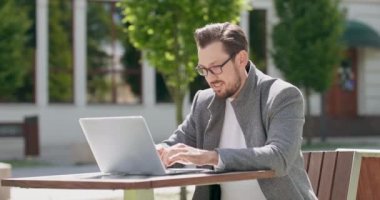 Gözlüklü, bıyıklı ve sakallı genç adam, meydandaki masada oturuyor, dizüstü bilgisayarında arka planda çalışıyor.
