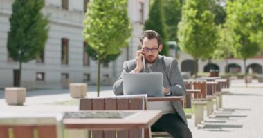 Gözlüklü, bıyıklı ve sakallı genç adam, meydanda masada oturuyor, dizüstü bilgisayarla çalışıyor ve arka plandaki telefon binalarıyla konuşuyor.