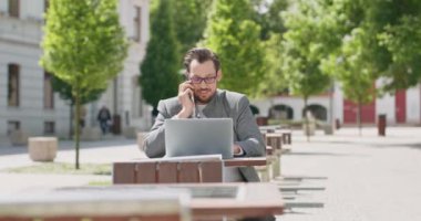 Gözlüklü, bıyıklı ve sakallı genç adam, meydanda masada oturuyor, dizüstü bilgisayarla çalışıyor ve arka plandaki telefon binalarıyla konuşuyor.