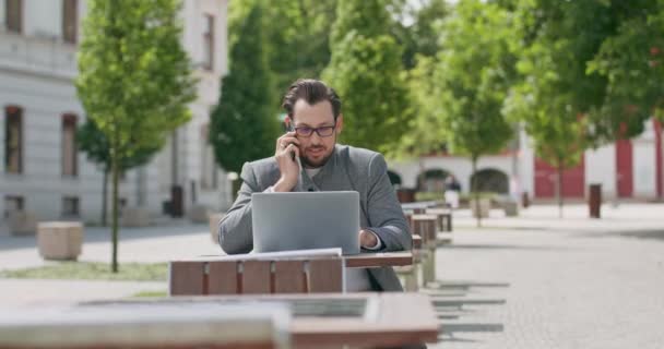 Jovem, vestindo óculos, com bigodes e barba, está sentado à mesa na praça, está trabalhando no laptop e falando ao telefone. Edifícios no fundo. Trabalhando fora — Vídeo de Stock