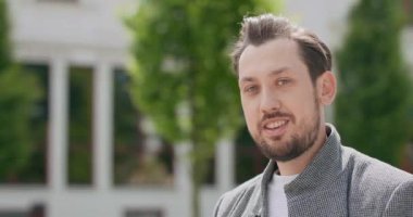 Bıyıklı ve sakallı genç adam, yüzünü kameraya doğru çeviriyor ve arka plandaki gülümseme binalarına doğru dönüyor.