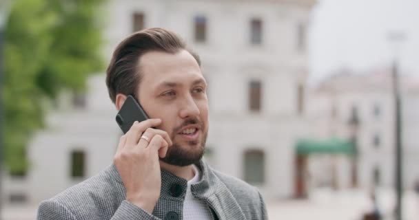 Joven, con bigotes y barba está hablando en el teléfono inteligente en la plaza Edificios en el fondo — Vídeos de Stock
