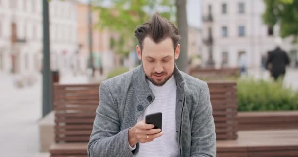 Joven con bigotes y una barba sentado en un banco en la plaza, desplazando el teléfono inteligente Edificios en el fondo — Vídeos de Stock