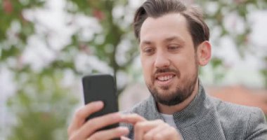 Bıyıklı, sakallı, meydanda oturmuş akıllı telefondan gülümseyen genç bir adamı yakalayın.