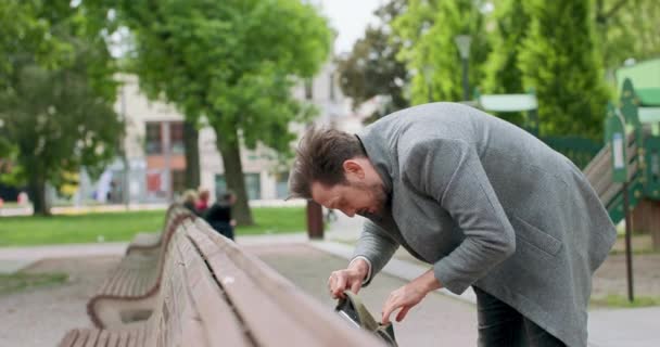 Молодой человек с усами и бородой ищет что-то в своем рюкзаке, помещенный на деревянной скамейке на площади Зеленые деревья на заднем плане — стоковое видео
