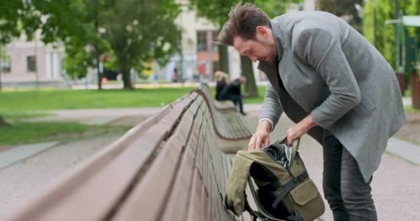 Junger Mann mit Schnurrbart und Bart sucht etwas in seinem Rucksack, der auf der Holzbank auf dem Platz abgestellt ist. Grüne Bäume sind im Hintergrund — Stockvideo