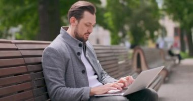 Bıyıklı ve sakallı genç bir adam meydanda bir bankta oturuyor, bankın arkasına yaslanıyor, dizüstü bilgisayarında daktilo yazıyor yeşil ağaçlar ve çimenler, binalar ve ahşap banklar
