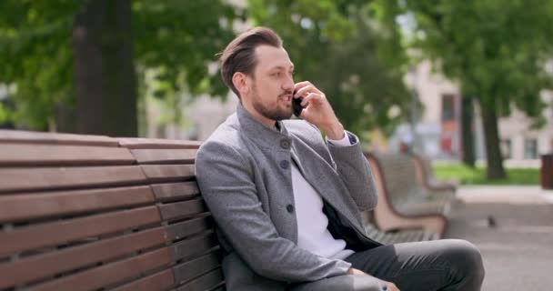 Jovem, com bigodes e barba está sentado em um banco, na praça, falando pelo smartphone Edifícios e árvores verdes no fundo — Vídeo de Stock