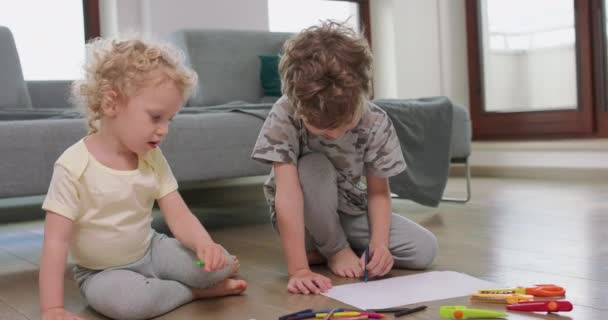 Mały chłopiec i mała dziewczynka rysują z ołówkami na białym papierze na podłodze patrząc na siebie od czasu do czasu Ołówki, papier, nożyczki dla dzieci i noże papierowe są przed nimi — Wideo stockowe
