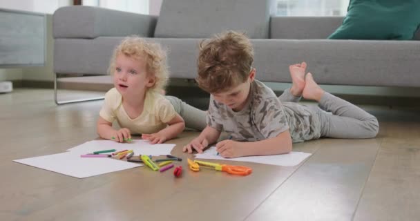 Un petit garçon et une petite fille sont allongés sur le sol et dessinent avec des crayons sur le papier blanc, et souriant Têtes levées à leurs parents et souriants Crayons, papier, enfants ciseaux et papier — Video