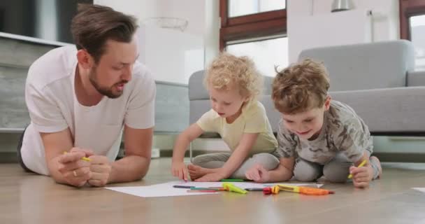 Ένα μικρό αγόρι και ένα μικρό κορίτσι, και ο πατέρας τους ζωγραφίζουν με μολύβια στο λευκό χαρτί στο πάτωμα. Ο πατέρας μιλάει με κορίτσια. Μολύβια, χαρτί, παιδικά ψαλίδια και χάρτινα μαχαίρια είναι μπροστά τους. — Αρχείο Βίντεο