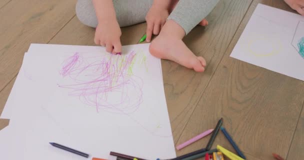 Közelről egy kislány kezét és lábát, aki ceruzával rajzol a fehér papírra a padlón ceruzák, papír van előtte. — Stock videók