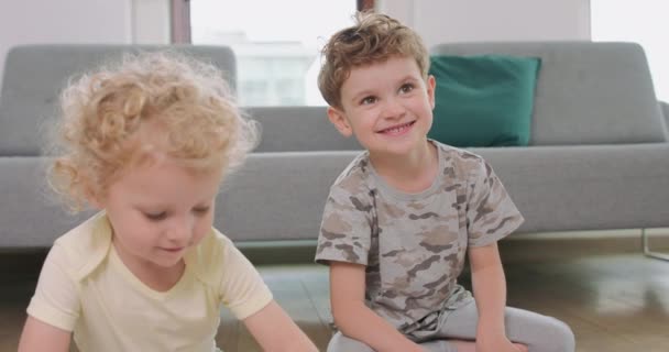 Großaufnahme eines kleinen Jungen und eines kleinen Mädchens, die auf dem Boden zeichnen Mädchen schaut auf ihre Zeichnung, Bruder blickt in die Kamera und lächelt Graues Sofa und Balkontür im Hintergrund — Stockvideo