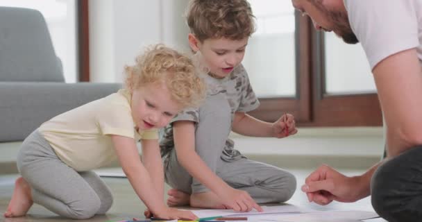 Közelkép egy kisfiúról és egy kislányról, és az apjuk ceruzákat rajzol a fehér papírra a padlón. Apa és a fiú a szürke kanapéról és az erkélyajtóról beszélnek. — Stock videók