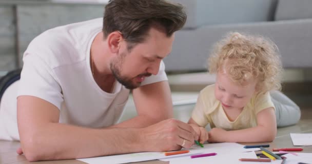 Gros plan d'une petite fille et de son père, qui dessinent sur le sol Père explique à sa fille quelque chose sur l'image Canapé gris en arrière-plan — Video