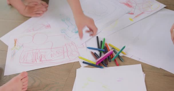 Närbild av en liten flickor och en liten pojkar gryningar, som ritar med pennor på vitt papper på golvet Pennor, papper är framför dem — Stockvideo