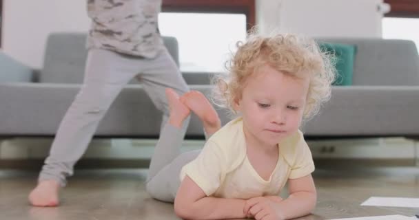Großaufnahme eines kleinen Mädchens, das auf dem Boden liegt Mädchen betrachtet ihre Zeichnung, Bruder springt hinter sie und sitzt lächelnd auf dem Boden Graues Sofa im Hintergrund — Stockvideo