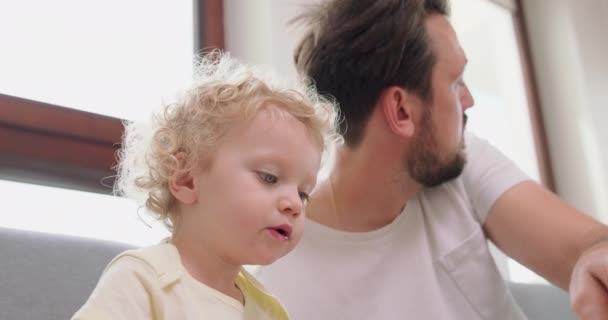 Closeup of a little girl and her bearded and mustashed father are looking pictures in book, sitting on gray sofa Father turnes for a while to the boy, who is sitting behind him — Stock Video