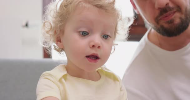 Closeup of a little girl and her bearded and mustashed father are looking pictures in book, sitting on gray sofa — Stock Video