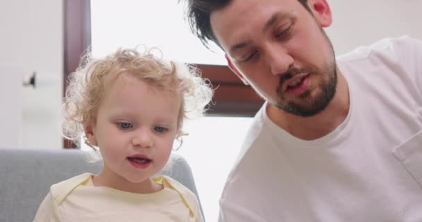 Close-up van een klein meisje en haar baard en mosterd vader zijn op zoek naar foto 's in boek, zittend op een grijze bank — Stockvideo