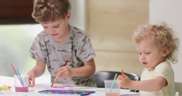 Irmão e irmã desenhar aquarelas enquanto sentado à mesa Menina tenta mergulhar pincel em aquarelas — Vídeo de Stock