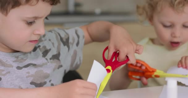 Hermano y hermana están cortando diferentes objetos de papel con tijeras, mientras están sentados a la mesa en la cocina — Vídeos de Stock