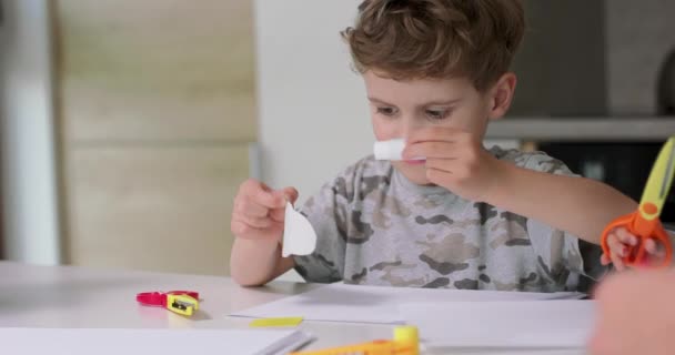 Close up de menino pequeno, que coloca a cola sobre o cortado de objetos de papel, durante a realização de aplicações com papel colorido e cola, enquanto sentado à mesa na cozinha Sua irmã corta algo — Vídeo de Stock