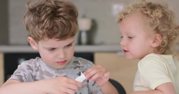 Close-up van kleine jongen, die de lijm op de gesneden van papieren voorwerpen, tijdens het maken van toepassingen met gekleurd papier en lijm, terwijl zitten met zijn zus aan de tafel in de keuken Zijn zus — Stockvideo