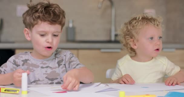 Kleiner Junge legt den Kleber auf die ausgeschnittenen Papierobjekte, während er mit farbigem Papier und Klebstoff Applikationen macht, während er mit seiner Schwester am Tisch in der Küche sitzt. — Stockvideo